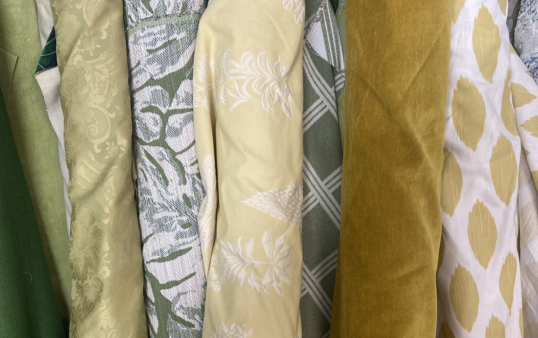 Stack of rolled designer fabric remnants in golden and green tones, including damask, botanical jacquard, plaid, velvet, and ikat dot patterns