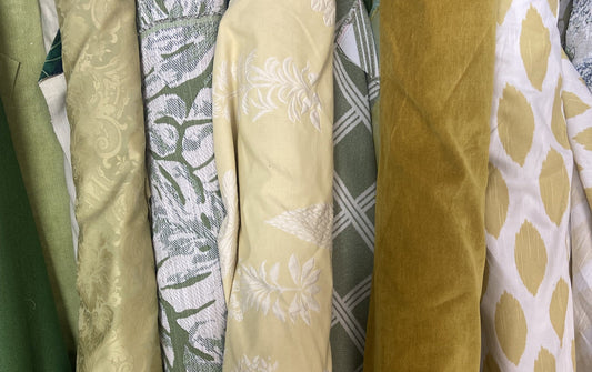 Stack of rolled designer fabric remnants in golden and green tones, including damask, botanical jacquard, plaid, velvet, and ikat dot patterns